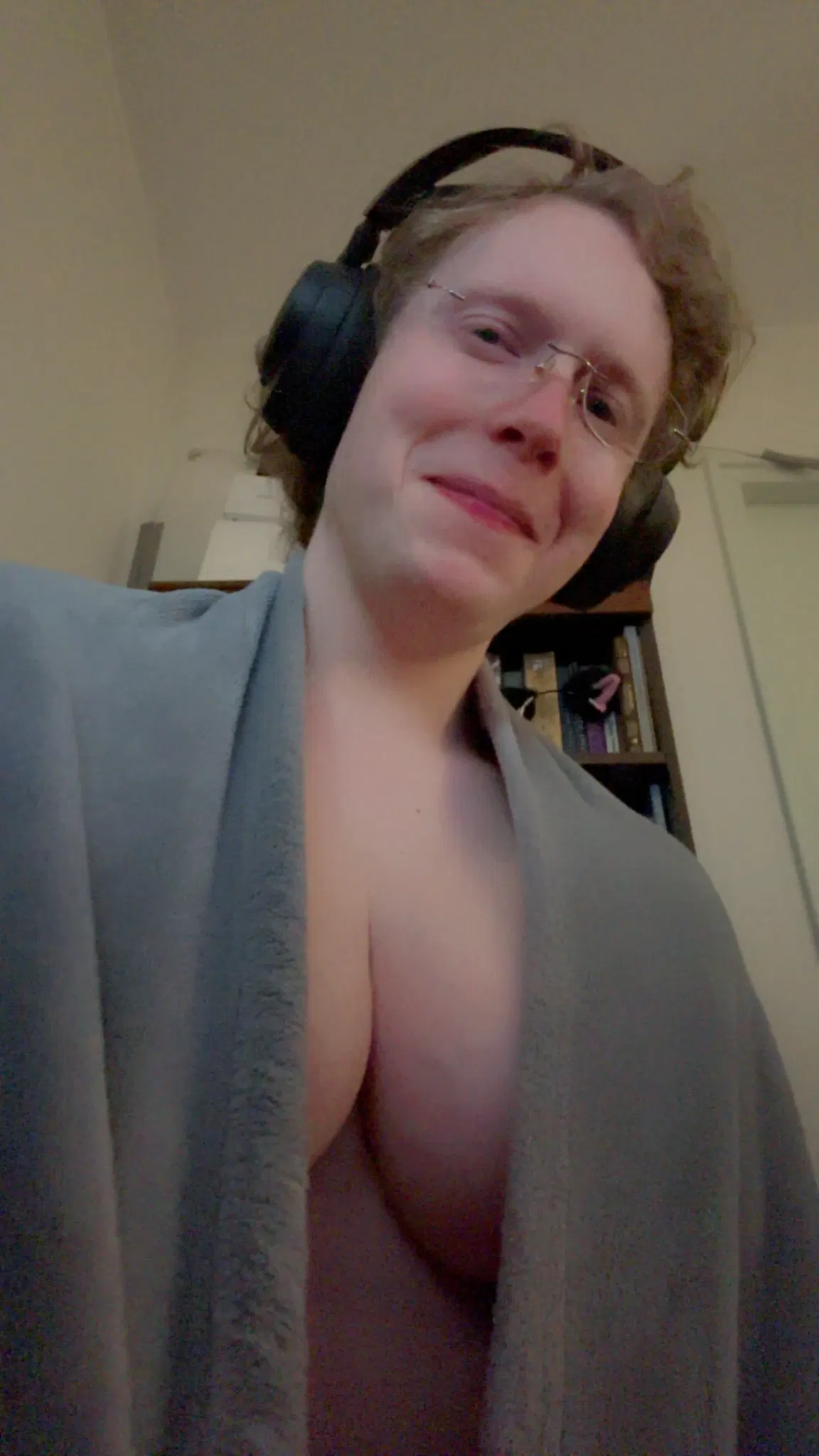 Siina in a grey blanket open at the center to show off maximum cleavage. She is looking down and smiling at the camera.